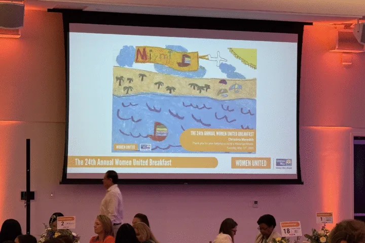 Colorful child’s drawing displayed on a large screen at the United Way Women United Breakfast, depicting a sunny Miami beach scene with an airplane banner, ocean waves, and a sailboat - presented as a gift to keynote speaker Christina Meredith.