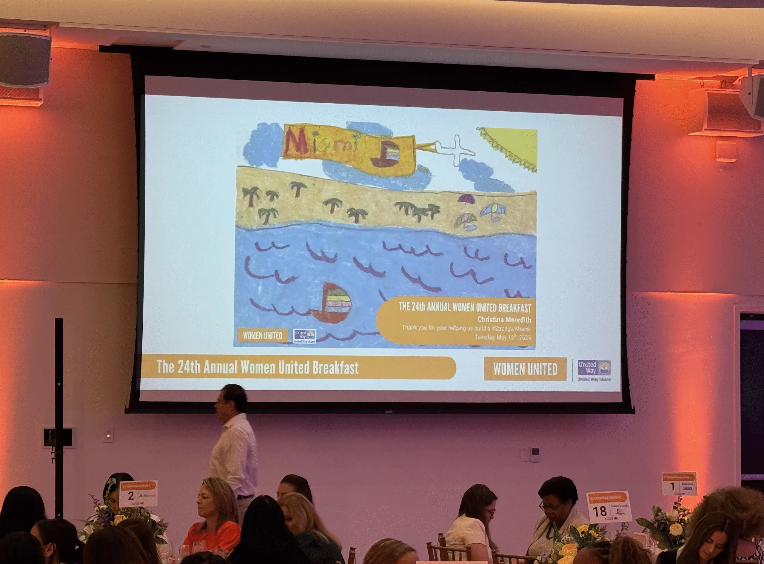 Colorful child’s drawing displayed on a large screen at the United Way Women United Breakfast, depicting a sunny Miami beach scene with an airplane banner, ocean waves, and a sailboat - presented as a gift to keynote speaker Christina Meredith.