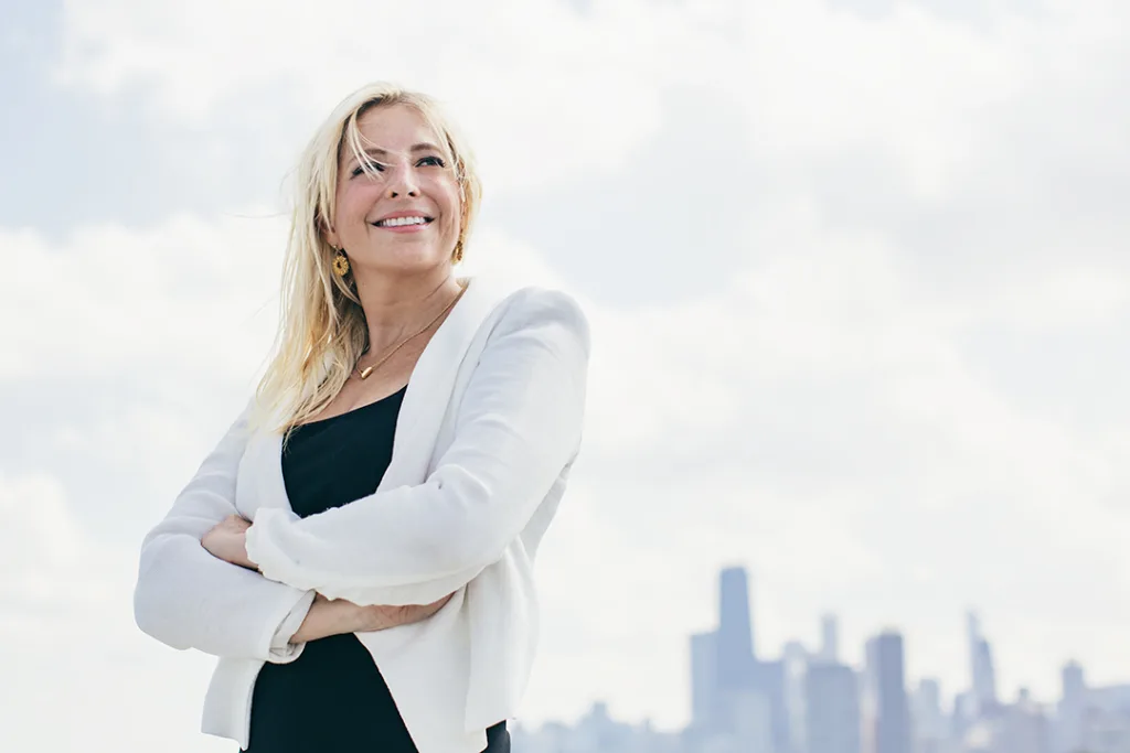 Aimee Baller smiling with arms crossed, standing confidently in front of a city skyline.